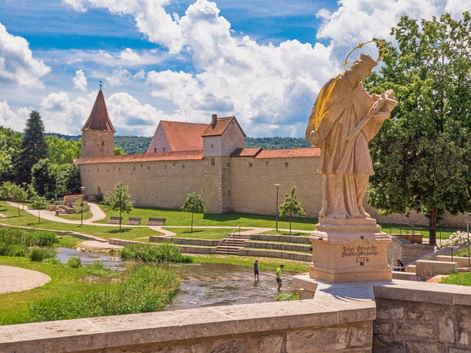 Steinstatue mit goldener Schärpe auf Brücke mit Blick auf flachen Bach in Berching. Mittelalterliche Stadtmauer mit Turm und roten Dächern hinter grünem Park.