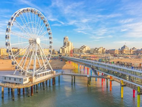 Scheveningen Strand mit weißem Riesenrad auf Pier und buntem Steg über dem Wasser. Sandstrand und Gebäude unter blauem Himmel sichtbar.