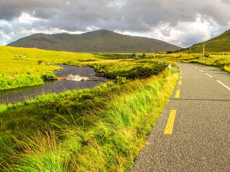 Asphaltstraße mit gelben Markierungen entlang des Bundorragha River durch grüne irische Landschaft im Delphi Valley mit Bergen unter bewölktem Himmel.
