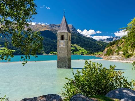 Historischer Kirchturm ragt aus dem türkisfarbenen Wasser des Reschensees mit Bergen im Hintergrund, eingerahmt von grünem Laub unter blauem Himmel.