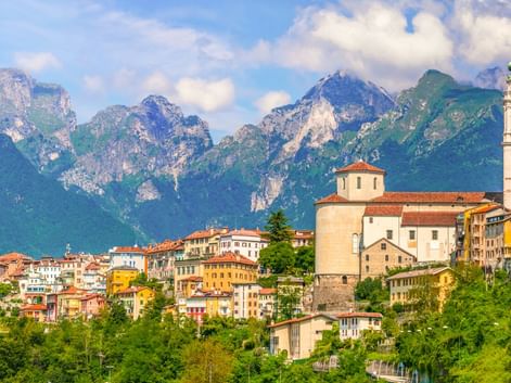 Historische Stadt Belluno mit bunten Gebäuden und einer Kirche mit Glockenturm vor dramatischen Berggipfeln unter blauem Himmel.