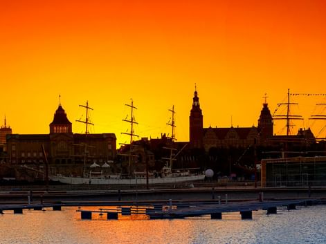 Silhouette der historischen Uferpromenade Stettins bei Sonnenuntergang mit Kirchtürmen und Segelschiffmasten vor orange-gelbem Himmel.