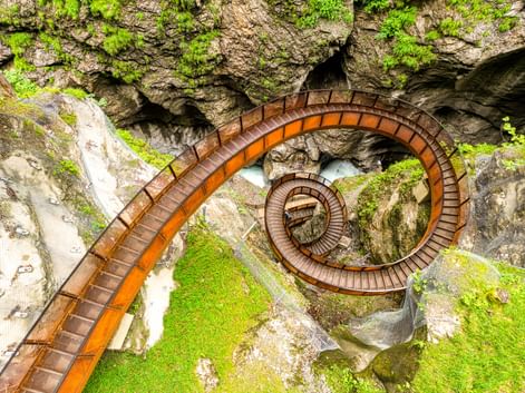 Luftaufnahme eines spiralförmigen Holzstegs in der Liechtensteinklamm mit moosbewachsenen Felswänden und fließendem Wasser.
