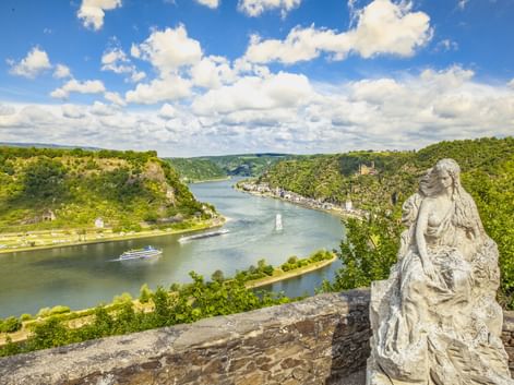 Steinerne Loreley-Statue in St. Goarshausen mit Blick auf den Rhein, der sich durch grüne Hügel schlängelt, mit einem Kreuzfahrtschiff auf dem Wasser.