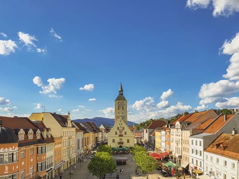 Historischer Stadtplatz in Deggendorf mit dem hohen Glockenturm des Alten Rathauses im Zentrum, umgeben von bunten Gebäuden und Bäumen.