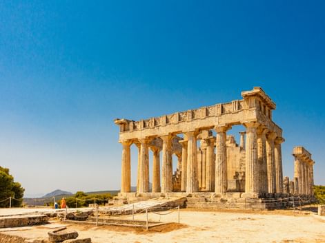 Antiker Aphaia-Tempel auf Ägina mit dorischen Säulen unter klarem blauen Himmel. Besucher erkunden die archäologische Stätte mit Bergen im Hintergrund.