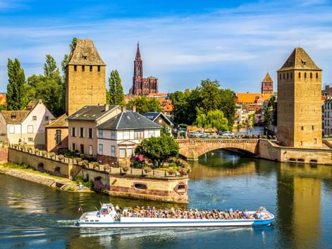 Touristenboot auf Fluss vor mittelalterlichen Türmen und Brücke in Straßburger Altstadt, mit Münsterspitze im Hintergrund unter blauem Himmel.