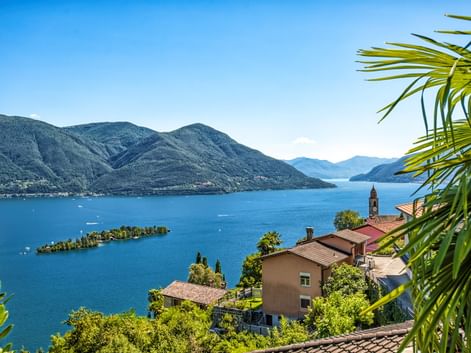 Panoramablick auf den Lago Maggiore mit kleinen Inseln, umgeben von Bergen. Dorfhäuser mit roten Dächern und Kirchturm im Vordergrund, Palmwedel sichtbar.