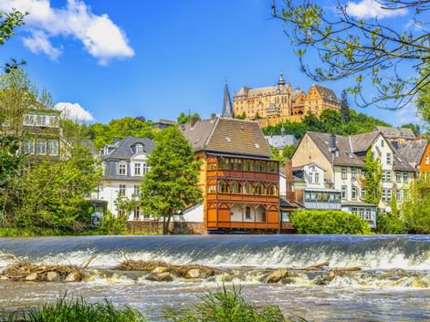 Blick auf Marburg mit Flusswehr im Vordergrund, historischen Fachwerkhäusern am Flussufer und dem Marburger Schloss auf dem Hügel.