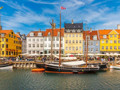 Historische Holzsegelschiffe am Nyhavn-Hafen in Kopenhagen mit bunten gelben, weißen und orangen Stadthäusern entlang der Uferpromenade.