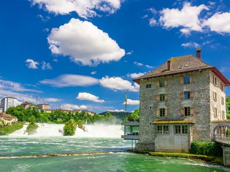 Rheinfall bei Schaffhausen mit kraftvoll strömendem Wasser. Historisches Steingebäude steht neben dem türkisfarbenen Fluss unter blauem Himmel.