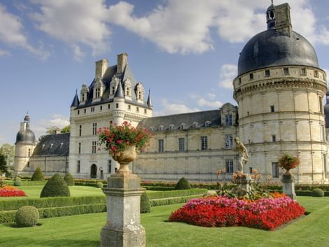 Französisches Renaissance-Schloss mit runden Türmen und Kuppeldächern. Gepflegte Gärten mit roten Blumen, geschnittenen Hecken und Steinurnen im Vordergrund.