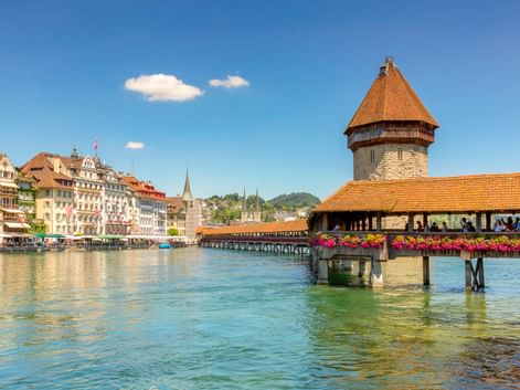 Historische Kapellbrücke mit orangem Dach und achteckigem Wasserturm über der Reuss in Luzern, mit bunten Gebäuden entlang des Ufers.