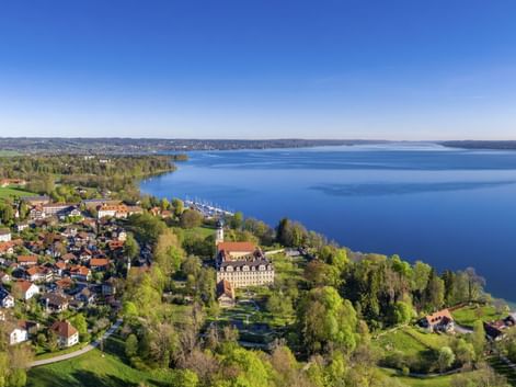 Luftaufnahme von Bernried am Starnberger See mit Dorf am Seeufer, rotgedeckten Häusern, großem historischen Gebäude und blauem Seewasser unter klarem Himmel.