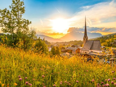Wallfahrtskirche Maria Alm mit hohem Turm im österreichischen Dorf bei Sonnenuntergang, umgeben von Wildblumenwiese und Bergen unter goldenem Himmel.