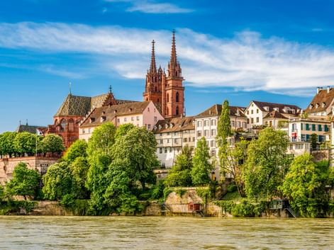 Blick auf die Basler Altstadt vom Rhein mit den beiden Türmen des roten Sandstein-Münsters über historischen Gebäuden und grünen Bäumen.