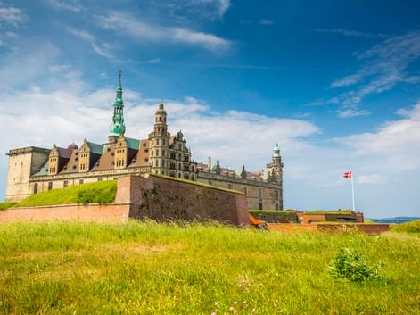 Schloss Kronborg in Helsingør mit grünen Kupfertürmen und Steinbefestigungen, umgeben von Grashängen. Dänische Flagge weht über den Mauern.