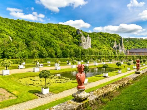 Formaler Garten in Freyr mit Zierteich, geschnittenen Bäumen, roten Statuen und dramatischen Kalksteinfelsen im Hintergrund unter blauem Himmel.