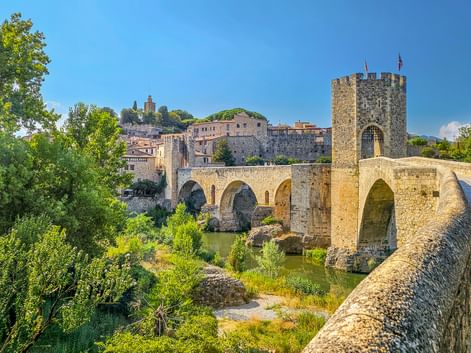 Mittelalterliche Steinbrücke mit Turm und Bögen über einem Fluss in Besalú, Spanien. Historische Gebäude und grüne Vegetation umgeben die Brücke.