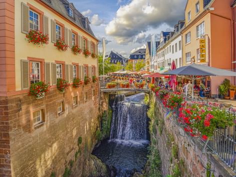 Wasserfall im historischen Zentrum von Saarburg, flankiert von bunten Gebäuden mit Blumenkästen und Straßencafés unter Sonnenschirmen.