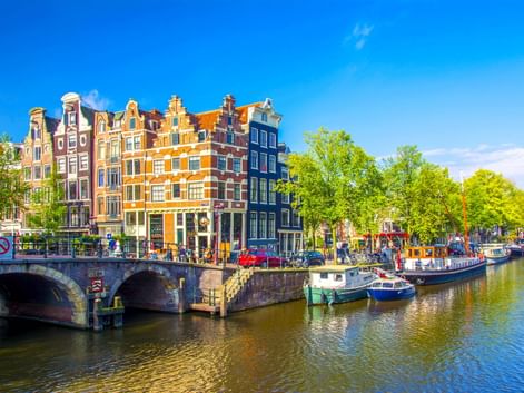 Amsterdamer Gracht mit traditionellen niederländischen Giebelhäusern in verschiedenen Farben, Booten am Ufer und Steinbrücke unter blauem Himmel.