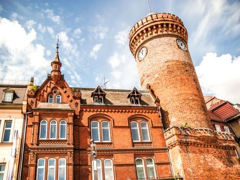Historischer Spremberger Turm in Cottbus mit Zifferblatt und Zinnen, neben rotem Backsteingebäude mit verzierten Giebeln unter blauem Himmel.