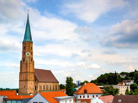 Spätgotische Hallenkirche St. Johannes in Dingolfing mit hohem Backsteinturm, grüner Spitze und Uhr, überragt rotgedeckte Häuser.