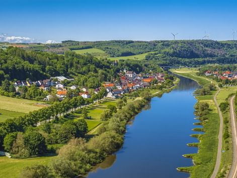 Aerial view of Beverungen with red-roofed houses along the Weser River, surrounded by green forests and hills. Wind turbines visible on distant hills.
