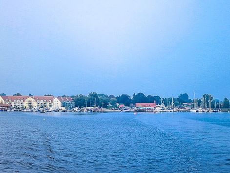 Blick über ruhiges blaues Wasser auf einen Hafen der Insel Poel mit Segelbooten, Wohngebäuden mit roten Dächern und Bäumen unter klarem Himmel.
