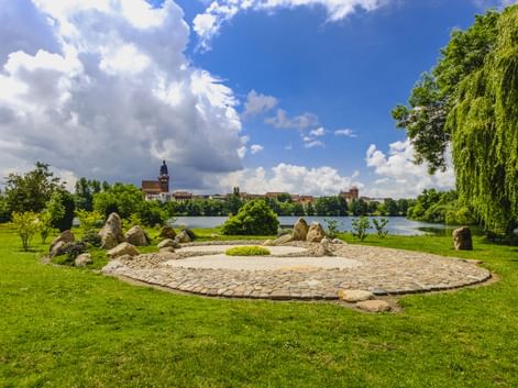 Kreisförmiger Steingarten mit großen Findlingen und Kies auf grüner Wiese am Tiefwarensee in Waren-Müritz. Kirchturm über dem See sichtbar.