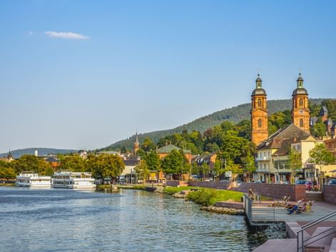 View of Miltenberg with St. Jakobus Church's twin towers along the Main River. Riverfront promenade with boats, hills in background.