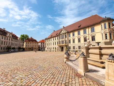 Kopfsteinpflasterplatz in Eichstätt mit verziertem Brunnen und gelben Barockpalais mit roten Ziegeldächern unter blauem Himmel.