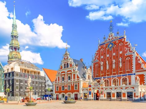 Das reich verzierte rote Schwarzhäupterhaus am Rathausplatz in Riga mit aufwendigen weißen Dekorationen und einer Uhr. Kirchturm links sichtbar.