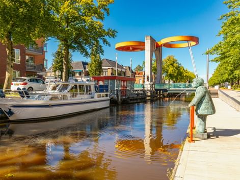 Bunte Eurobrug-Zugbrücke mit gelb-roten Scheiben über Kanal in Stadskanaal. Boote am Ufer, grüne Bäume und Statue sichtbar.
