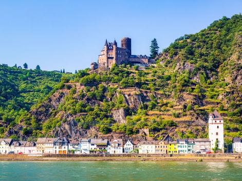 View of St. Goar on the Rhine with medieval castle on terraced vineyard hillside. Colorful buildings line the riverbank below steep slopes.