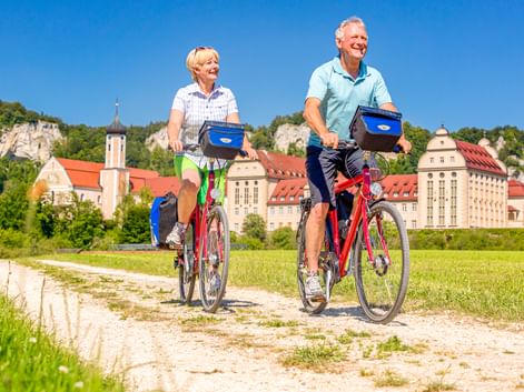 Two cyclists on red bikes riding on a gravel path with Beuron Monastery and limestone cliffs in the background under clear blue sky.