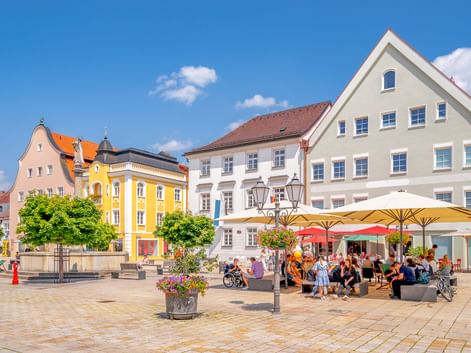Sonniger Marktplatz in Schongaus Altstadt mit bunten historischen Gebäuden, Straßencafé mit Sonnenschirmen, Brunnen und Menschen.