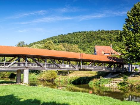 Überdachte Holzbrücke mit rotem Dach über einem ruhigen Fluss in Fridingen. Grüne Wiese im Vordergrund, bewaldete Hügel und Häuser im Hintergrund.