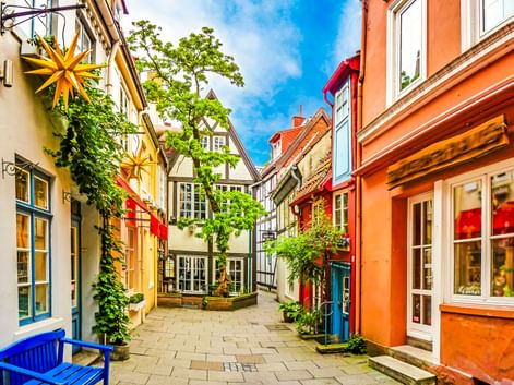 Schmale Kopfsteinpflasterstraße in Bremen, gesäumt von bunten Fachwerkhäusern in Gelb, Orange und Rot. Grüne Pflanzen und Sterne schmücken die Fassaden.