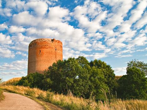 Round brick tower on a hill surrounded by green trees and grass under a blue sky with white clouds. A path leads toward the structure.
