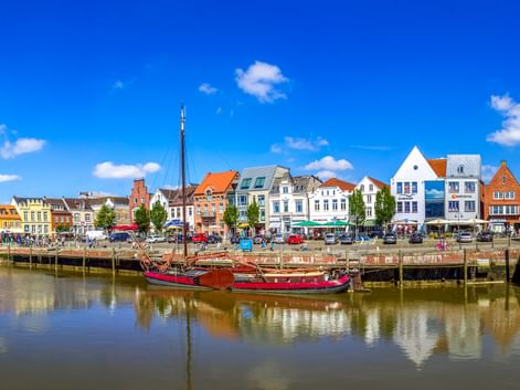 Historisches hölzernes Segelboot am Husumer Hafen mit bunten traditionellen Gebäuden entlang der Uferpromenade unter blauem Himmel.