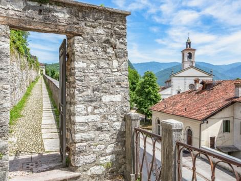 Alter Steinbogen mit Holztor entlang eines schmalen Weges in Feltre, mit weißem Kirchturm und Bergen im Hintergrund.