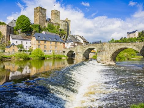 Mittelalterliche Burg auf einem Hügel über einer Steinbogenbrücke, die einen Fluss mit Wehr in Runkel überspannt. Bunte Häuser säumen das Flussufer.