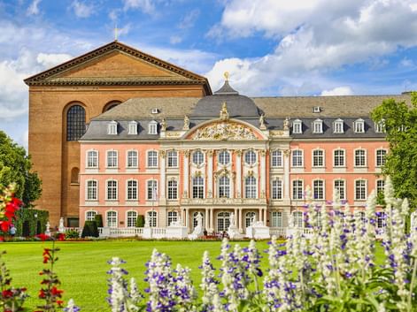 Kurfürstliches Palais mit rosa Rokoko-Fassade und Konstantinbasilika in Trier. Bunte Blumenbeete blühen im Palastgarten unter blauem Himmel.