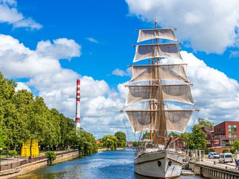 Großes Segelschiff mit weißen Segeln am Dane-Fluss in Klaipeda, Litauen. Grüne Bäume säumen den Wasserweg unter blauem Himmel mit Wolken.