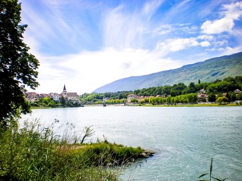 View across the Rhône River in Seyssel, France, showing a church tower and historic buildings along the waterfront with green mountains in the background.