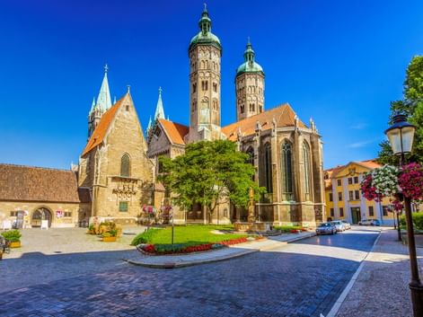 Naumburger Dom mit zwei hohen Türmen und orangefarbenen Dächern auf einem sonnigen Platz. Blumenbeete und Bäume schmücken den Kopfsteinpflasterplatz.