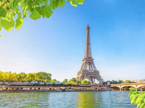 Eiffelturm über der Seine in Paris an einem sonnigen Tag, eingerahmt von grünen Blättern. Eine Brücke und baumgesäumtes Flussufer sind sichtbar.