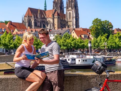 Two cyclists with map at Danube riverfront, red bicycle nearby. Regensburg Cathedral with twin spires rises above red-roofed buildings.