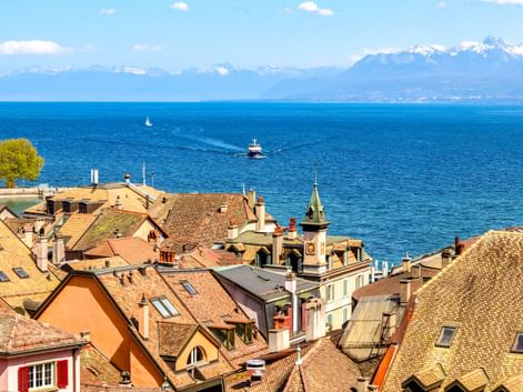 Traditionelle Ziegeldächer von Nyon mit Genfersee und schneebedeckten Alpen im Hintergrund. Boote fahren auf dem blauen Wasser unter klarem Himmel.